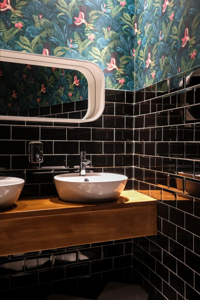 Modern bathroom interior with double sinks, black tile walls, wooden counter, and tropical wallpaper, creating a stylish and elegant atmosphere.