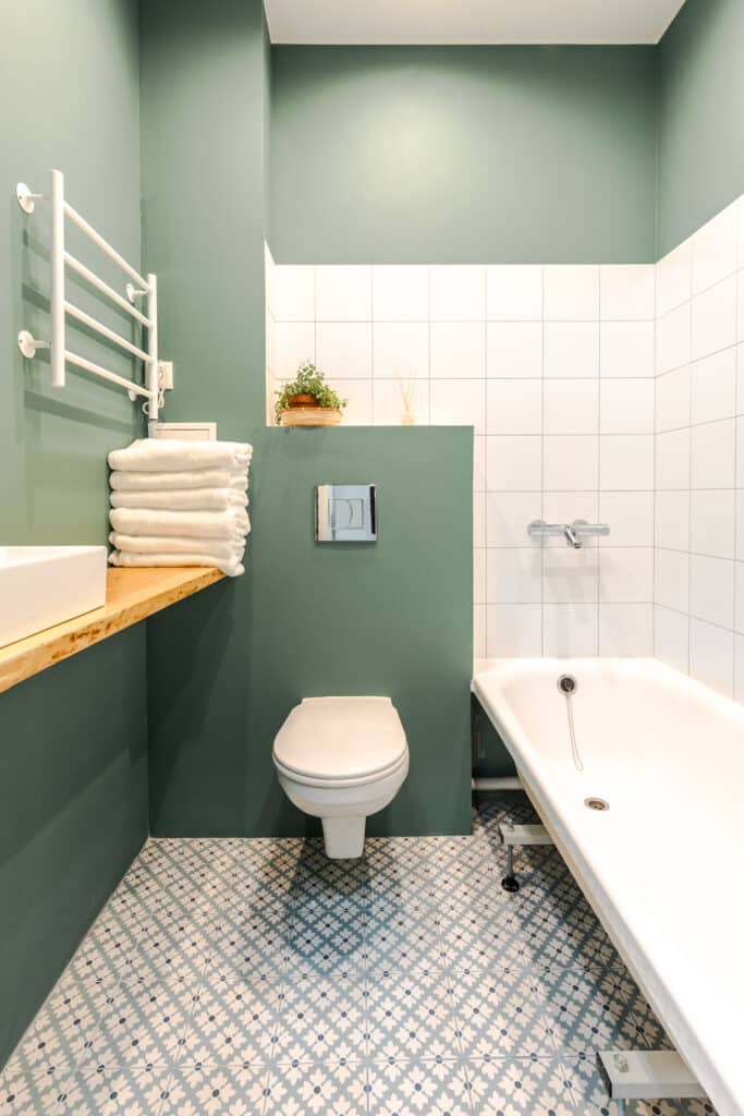 Salle de Bain Moderne Vert Sauge et Carrelage Fleuri Salle de bain moderne avec murs vert sauge, carrelage au sol fleuri bleu et blanc, WC, baignoire et porte-serviettes blanc.