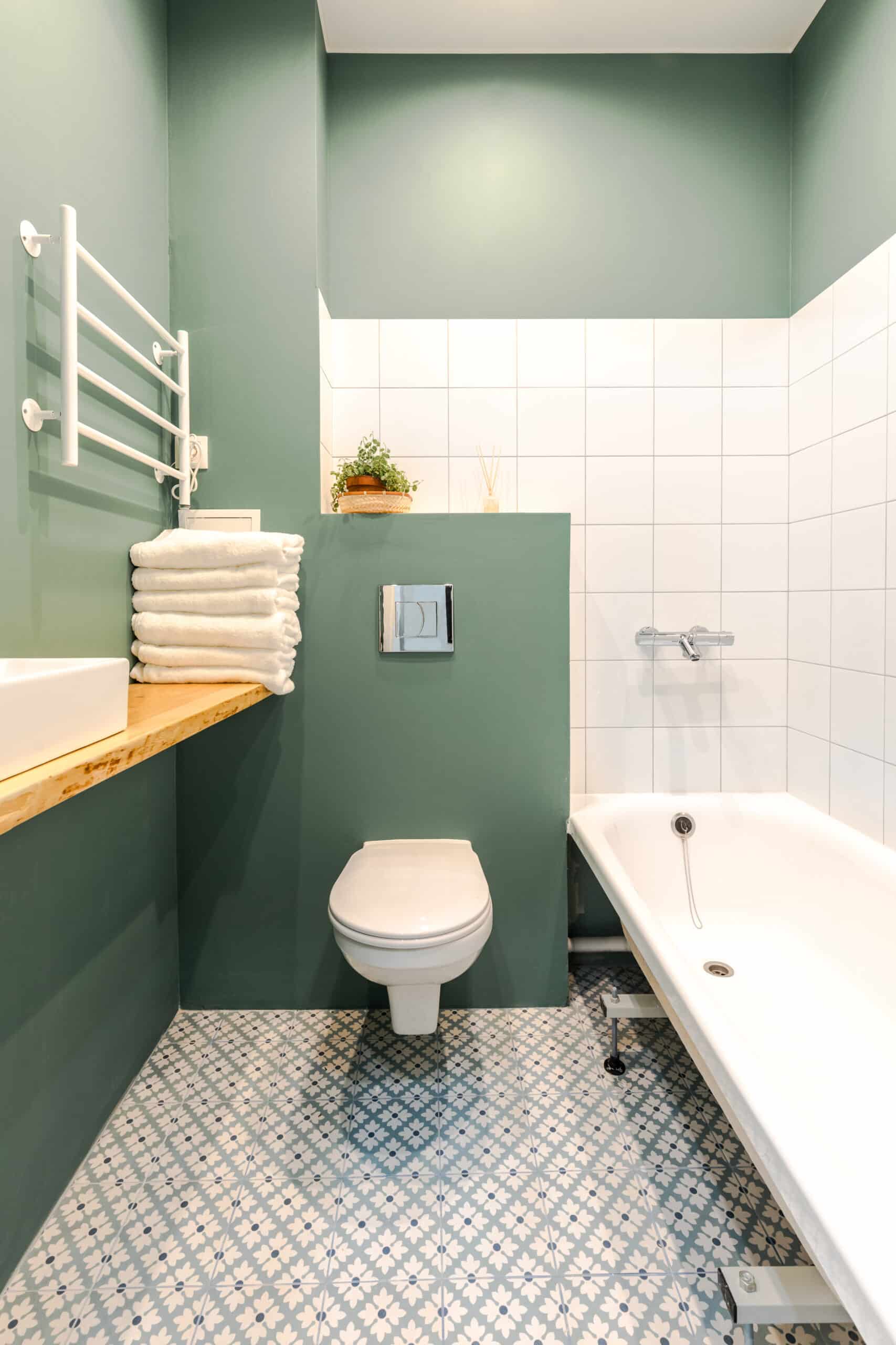 Salle de bain moderne avec murs vert sauge, carrelage au sol fleuri bleu et blanc, WC, baignoire et porte-serviettes blanc.